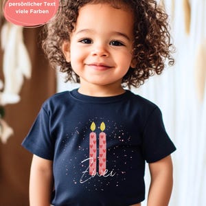 May include: A navy blue t-shirt featuring a design of two pink candles with heart details and yellow flames. The word "Zwei" is written in white script below the candles. The shirt is worn by a smiling child.