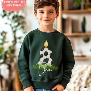 Könnte beinhalten: Ein dunkelgrünes Langarm-Sweatshirt mit einer Grafik der Zahl Acht im Design eines Fußballs mit einer brennenden Kerze darauf, platziert über dem weißen, kursiven Schriftzug Ben auf einem grünen Grasbüschel.