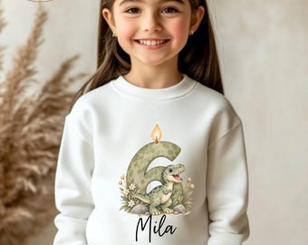 May include: A white long-sleeve sweatshirt featuring a graphic of a green dinosaur standing in front of a large number six shaped like a candle with a lit flame. The name Mila is printed in black script below the dinosaur graphic.