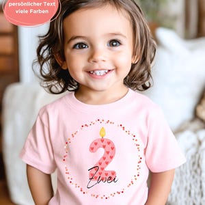 May include: A light pink t-shirt featuring a large number "2" in a candle design, decorated with red hearts. The word "Zwei" is written below the number. The shirt is encircled by a dotted border and worn by a smiling child.