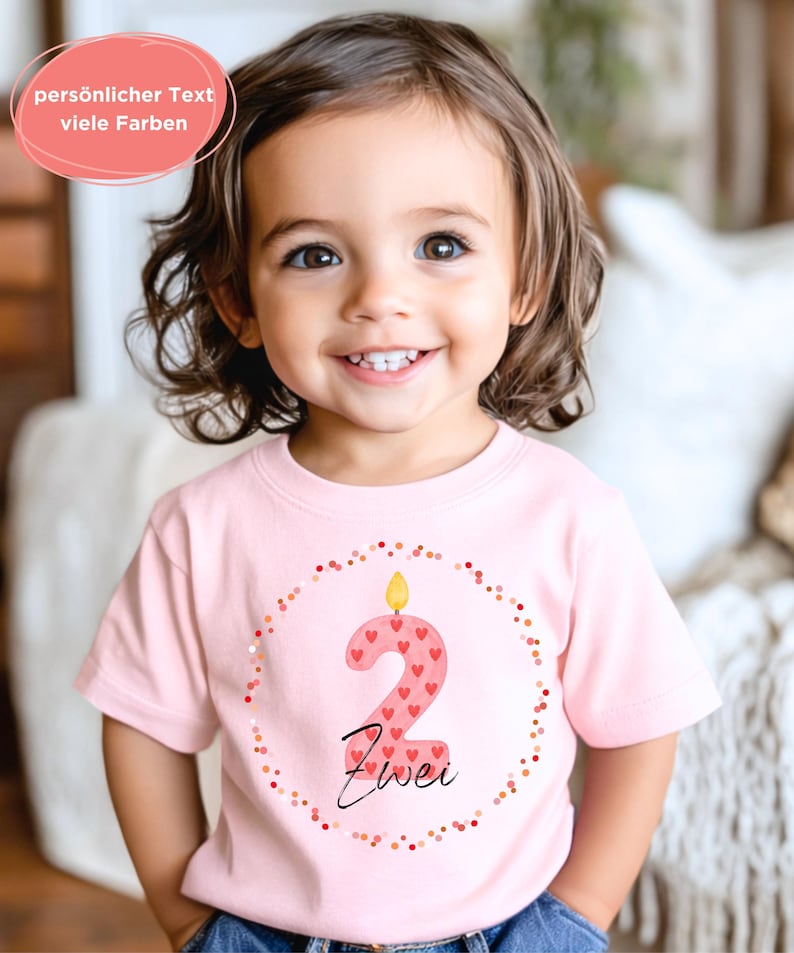 May include: A light pink t-shirt featuring a large number "2" in a candle design, decorated with red hearts. The word "Zwei" is written below the number. The shirt is encircled by a dotted border and worn by a smiling child.