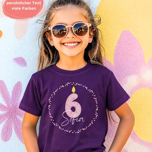 Könnte beinhalten: Ein dunkelviolettes Kurzarm-T-Shirt mit einer Grafik einer rosafarbenen Kerze in Form der Zahl sechs mit bunten Punkten, umgeben von einem kreisförmigen Konfettirand, mit dem Namen Sofia in weißer Schreibschrift unter der Kerze.
