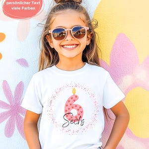 K&ouml;nnte beinhalten: Ein wei&szlig;es T-Shirt mit einem rosa Kerzenmotiv mit der Zahl 6, Herzd details und dem Wort "Sechs". Das T-Shirt wird von einer jungen Person mit Sonnenbrille getragen.
