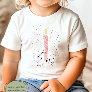 May include: White t-shirt featuring a pink birthday candle with a yellow flame and the word "Eins" in black script. The shirt has colorful confetti and is worn by a child.