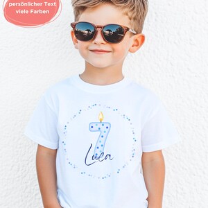 K&ouml;nnte beinhalten: Ein wei&szlig;es Kurzarm-T-Shirt mit einer Grafik einer hellblauen Kerze in Form der Zahl sieben mit brennender Flamme, umgeben von einem Kreis aus kleinen blauen Punkten und dem Namen Luca in dunkelblauer Schreibschrift unter der Kerze.