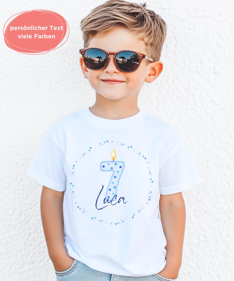 K&ouml;nnte beinhalten: Ein wei&szlig;es Kurzarm-T-Shirt mit einer Grafik einer hellblauen Kerze in Form der Zahl sieben mit brennender Flamme, umgeben von einem Kreis aus kleinen blauen Punkten und dem Namen Luca in dunkelblauer Schreibschrift unter der Kerze.