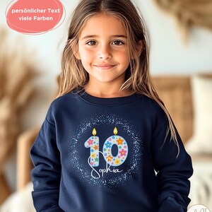 Könnte beinhalten: Ein dunkelblaues Langarm-Sweatshirt mit einer Grafik der Zahl 10, gestaltet als zwei hellblaue Kerzen mit bunten Blumenmustern und brennenden Flammen, umgeben von einem weiß gesprenkelten Kreis und dem Namen Sophia in weißer Schreibschrift.