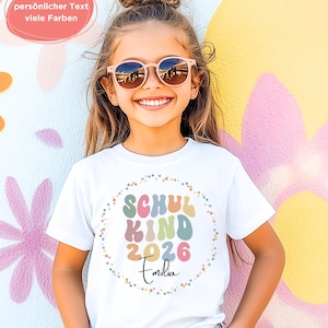 May include: A white t-shirt with the text "SCHUL KIND 2026 Emilia" in colourful, rounded letters. The text is encircled by a dotted border. The image also includes the text "personlicher Text viele Farben" in a pink bubble.