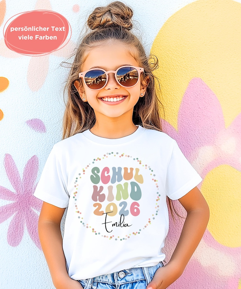 May include: A white t-shirt with the text "SCHUL KIND 2026 Emilia" in colourful, rounded letters. The text is encircled by a dotted border. The image also includes the text "personlicher Text viele Farben" in a pink bubble.