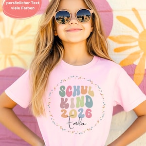 May include: A young person wearing a light pink t-shirt with the words "SCHUL KIND 2026 Emilia" in a colourful design. The person is wearing round sunglasses and smiling. The background features a colourful mural.