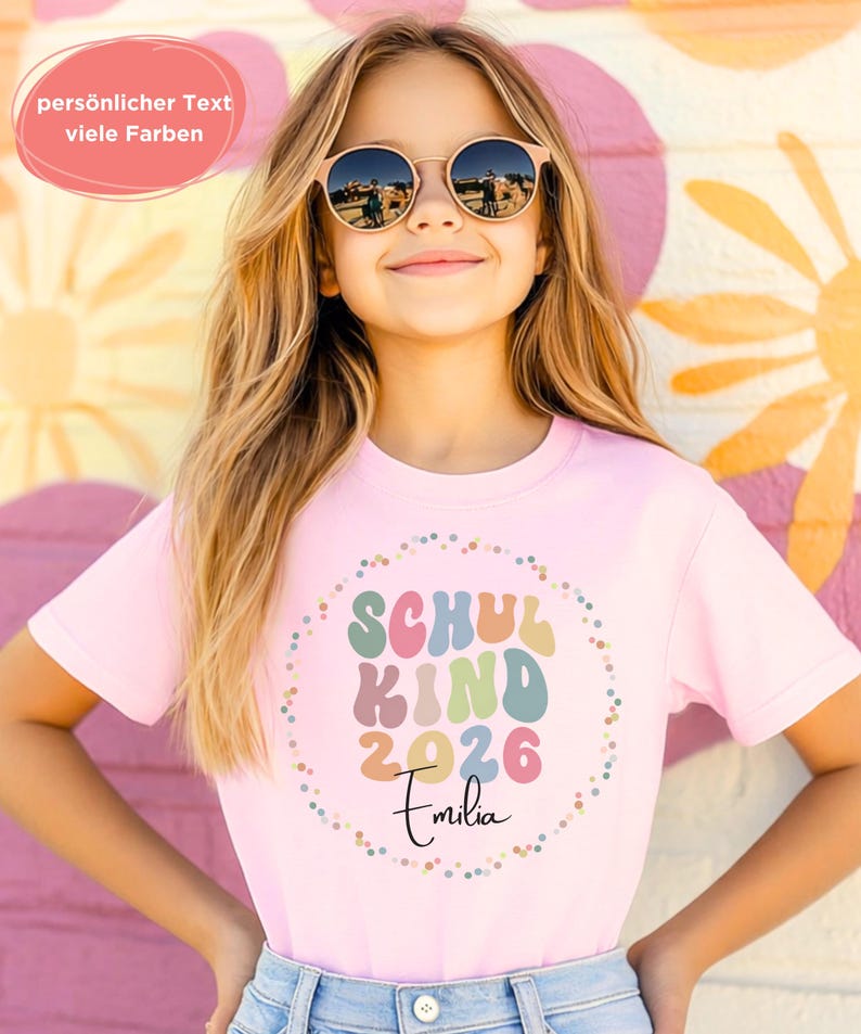 May include: A young person wearing a light pink t-shirt with the words "SCHUL KIND 2026 Emilia" in a colourful design. The person is wearing round sunglasses and smiling. The background features a colourful mural.