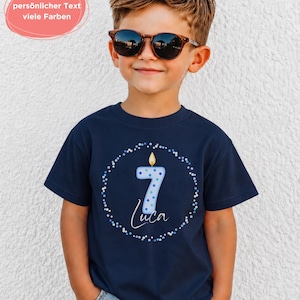 K&ouml;nnte beinhalten: Ein dunkelblaues Kurzarm-T-Shirt mit einer Grafik einer hellblauen Kerze in Form der Zahl sieben mit brennender Flamme, umgeben von einem kreisf&ouml;rmigen Rand aus blauen und wei&szlig;en Punkten sowie dem Namen Luca in wei&szlig;er Schreibschrift.