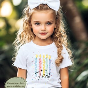 May include: A young girl with blonde curly hair and a white bow headband wears a white t-shirt. The shirt features a colorful design of birthday candles and the word "F&uuml;nf". The background is a blurred outdoor scene.