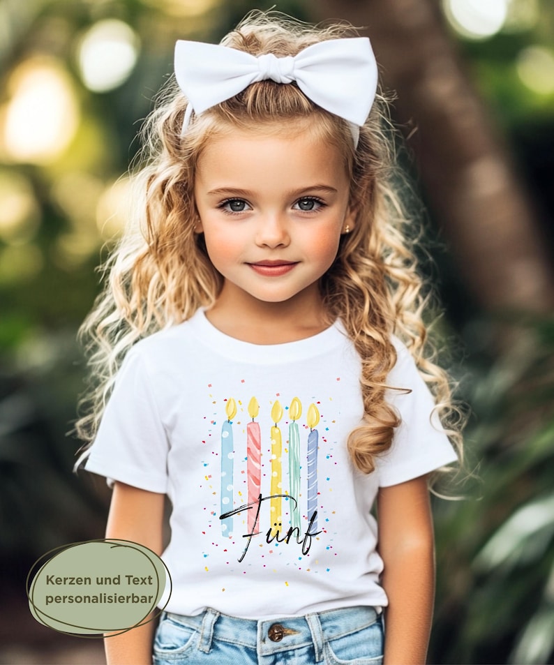 May include: A young girl with blonde curly hair and a white bow headband wears a white t-shirt. The shirt features a colorful design of birthday candles and the word "F&uuml;nf". The background is a blurred outdoor scene.