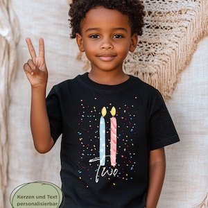 May include: A young child wearing a black t-shirt with a birthday theme. The shirt features two stylized candles, one blue and one pink, with the word "two" in white script. Colorful confetti dots the shirt. The child is smiling and holding up two fingers.