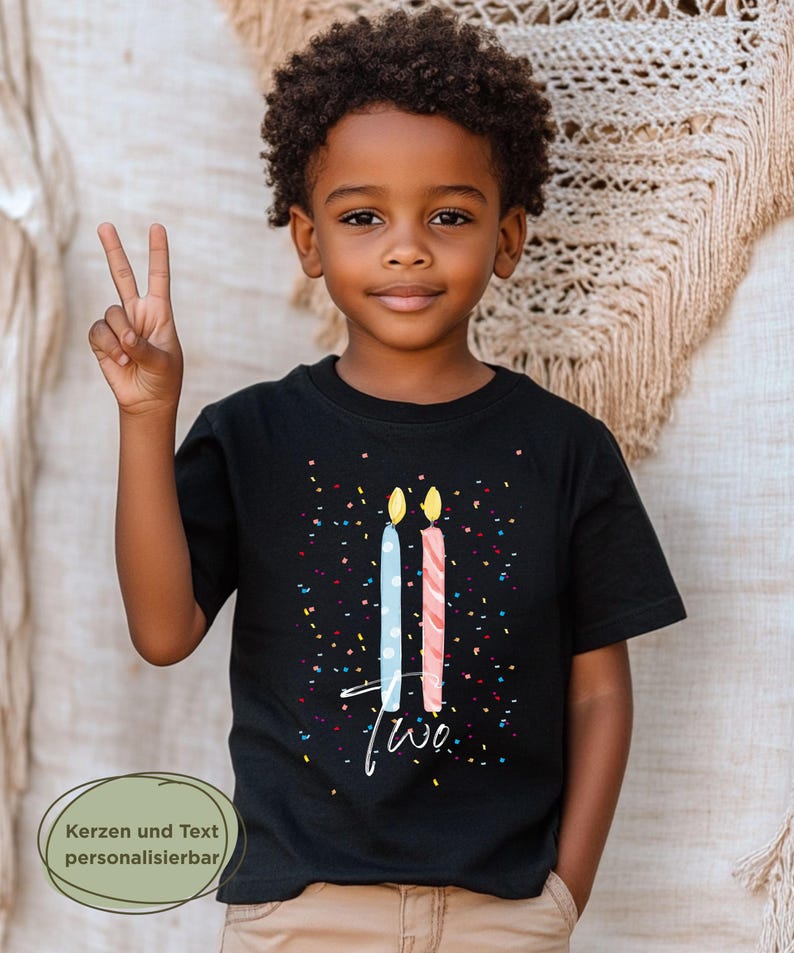 May include: A young child wearing a black t-shirt with a birthday theme. The shirt features two stylized candles, one blue and one pink, with the word "two" in white script. Colorful confetti dots the shirt. The child is smiling and holding up two fingers.