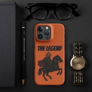 Puede incluir: Funda de teléfono naranja con un diseño del Jinete sin Cabeza montado en un caballo negro, con el texto "THE LEGEND". La funda está junto a un reloj dorado, gafas negras y un lápiz.