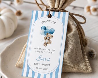May include: A small burlap bag tied with twine, featuring a blue and white striped tag that reads "Thank You" and "Eva's Baby Shower." The tag has a teddy bear illustration with balloons. The bag sits on a speckled ceramic plate, with decorative pumpkins in the background.