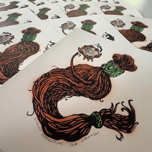 May include: A print on off-white paper featuring a stylized illustration of a long, wavy brown hair lock tied with green bands. The hair holds a small, detailed circular object. The text 'RINGLET REFLECTION 1/20 C VAMMEN' is printed at the bottom.