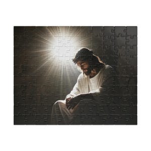 Op de afbeelding: Een rechthoekige legpuzzel met een afbeelding van een figuur met lang haar en een doornenkroon, gekleed in een gewaad, zittend met het hoofd gebogen. Een helder, stralend licht schijnt van achter de figuur tegen een donkere achtergrond.