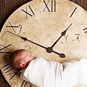 以下が含まれることがあります： 白い布に包まれた新生児が、アンティーク風の大きな時計の文字盤の上にいます。時計はローマ数字で、暗い色の針が2時頃を指しています。時計の文字盤は、質感があり、古びた外観をしています。