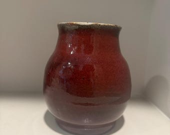 Könnte beinhalten: Eine kleine, handgefertigte Keramikvase mit tiefroter Glasur. Die Vase hat einen abgerundeten Körper, einen leicht ausgestellten Rand und ein dunkelbraunes Band um den oberen Rand. Die Vase ist etwa 10 cm hoch und 8 cm breit, geeignet für kleine Blumengestecke.