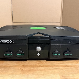 Puede incluir: Una consola de videojuegos Xbox original de color negro con un diseño superior en forma de X y un logotipo circular verde en el centro. El panel frontal muestra el logotipo de Xbox, una bandeja de disco, un botón de encendido y cuatro puertos.
