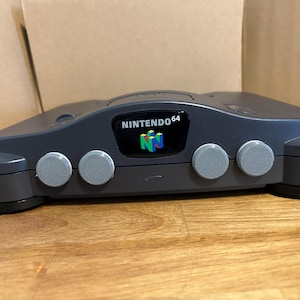 Puede incluir: Una consola de juegos Nintendo 64 de color gris oscuro. La consola tiene cuatro botones circulares grises en la parte delantera y el logotipo de Nintendo 64. La consola está sobre una superficie de madera.