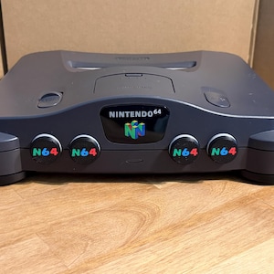 May include: A dark gray Nintendo 64 video game console featuring a central black logo plate with the text NINTENDO 64 and a colorful N64 logo. The front panel includes four controller ports, each covered by a black circular cap with N64 printed in green, blue, and red.