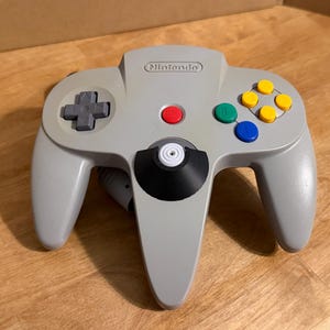 May include: A grey Nintendo 64 video game controller featuring a central analogue stick with a black base, a grey directional pad, a red start button, and four coloured action buttons in yellow, green, and blue, with the Nintendo logo embossed at the top.