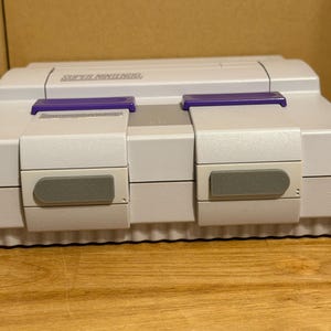 May include: A light gray Super Nintendo video game console featuring two purple controller ports on the front panel. The device has a rectangular shape with a top-loading cartridge slot and two gray rectangular buttons located below the controller ports.