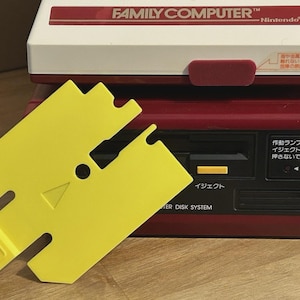 Op de afbeelding: Een rood-witte Nintendo Family Computer Disk System console met een gele plastic schijfstationbeschermer. De console heeft een zwart voorpaneel met Japanse tekst, een gele uitwerpknop en een rood-wit bovengedeelte met de tekst FAMILY COMPUTER.