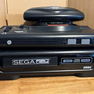 Puede incluir: Una consola Sega Genesis negra con un accesorio Sega CD. La consola tiene botones de encendido, volumen y reinicio. La unidad Sega CD tiene un indicador de acceso al disco y el logotipo de Sega.