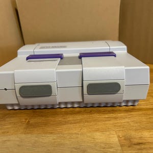 May include: A light gray Super Nintendo Entertainment System console. The console has a rectangular shape with a series of buttons and a slot for game cartridges. It features purple accents and gray buttons. The console is on a wooden surface.