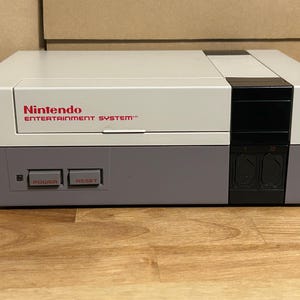 May include: A Nintendo Entertainment System console in a light gray and purple color scheme. The console has the Nintendo logo in red, with "POWER" and "RESET" buttons. The console is on a wooden surface.