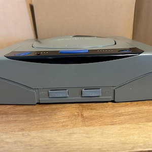 May include: A gray Sega Saturn video game console. The console has a rounded top with a disc tray and a black and blue control panel. The console is on a wooden surface.