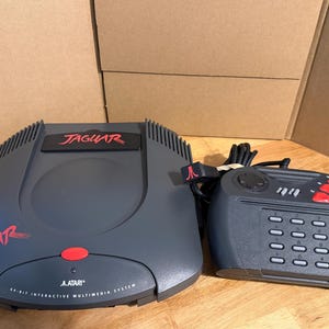 May include: A dark grey Atari Jaguar 64-bit interactive multimedia system console with a red power button and red Jaguar logo. Beside it is a matching dark grey controller featuring a numeric keypad, a directional pad, and three red action buttons.