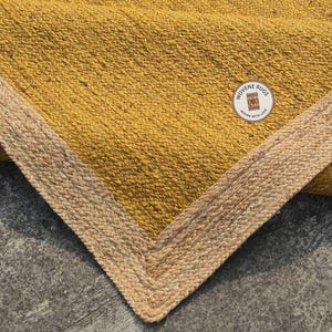 May include: A close-up of a mustard yellow rug with a woven texture. The rug has a natural fiber border in a lighter tan color. The rug is folded over, showing a corner of the rug. The rug has a tag that says "Woven Rugs, Woven with Love."
