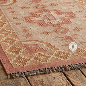 May include: A rectangular rug with a geometric pattern in shades of peach, tan, and brown. The rug has a fringed edge and is displayed on a wooden surface. The design features repeating shapes and a central medallion.