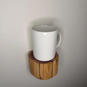 May include: A white ceramic mug rests on a small, natural wooden shelf. The shelf is made from a section of a tree trunk, displaying the wood grain. The mug has a simple, cylindrical shape with a handle.
