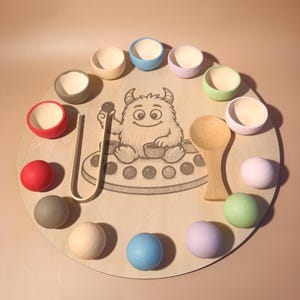 Op de afbeelding: Een houten educatief speelgoed met een cirkelvormig bord met een monsterillustratie. Het bevat gekleurde kommen, houten ballen, een houten lepel en een tang om te sorteren. Het bord heeft een diameter van ongeveer 30 cm.