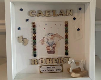 Op de afbeelding: Een ingelijste babyherinnering met de namen "CAELAN" en "ROBERT" in houten letters. De lijst bevat een afbeelding van een luchtballon, knopen, steraccenten, babyvoetjes en een houten schommelpaard. De lijst bevat ook het geboortegewicht en de datum van de baby.