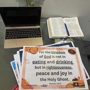 May include: A gold laptop, an open Bible with a blue and red pen, and several printed sheets with text and food illustrations are on a glass table. The text on the sheets reads "For the kingdom of God is not in eating and drinking but in righteousness, peace and joy in the Holy Ghost."