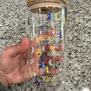 May include: A clear glass tumbler with a bamboo lid. The tumbler is decorated with blue and yellow graphics, including the words "Amazing" and "Kind", along with handprints, socks, and the numbers "321".