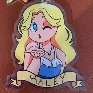 Peut inclure: Un porte-clés en acrylique transparent représentant une illustration de personnage blond aux yeux bleus, portant un haut bleu et un collier, envoyant un baiser. Une bannière jaune en bas affiche le nom HALEY en noir. Avec fermoir étoile doré.