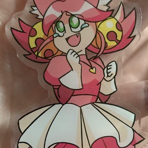 May include: A clear acrylic charm featuring a cartoon character with pink hair, green eyes, and a single visible fang. The character wears a pink dress with a white skirt, a yellow heart pendant, and has yellow circular accents in their hair.