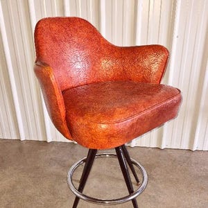 May include: A vintage orange bar stool with a textured seat and backrest. The stool has a chrome footrest and black legs with gold-colored tips. The chair is a retro design.