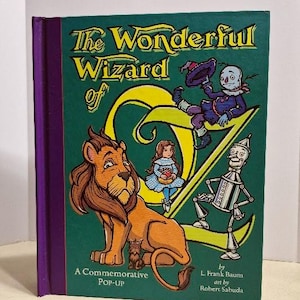 May include: A hardcover book titled "The Wonderful Wizard of Oz" with a green cover and purple spine. The cover features illustrations of the Lion, Dorothy, Scarecrow, Tin Man, and the Wicked Witch. Text includes "A Commemorative POP-UP" and author and artist credits.
