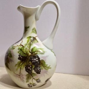 May include: A white ceramic pitcher with a curved handle and a spout, decorated with a blackberry and leaf design. A matching white stopper is next to the pitcher. The pitcher is likely for serving beverages.