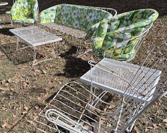 May include: Outdoor patio furniture set with white metal frames. Includes a loveseat, two armchairs, two side tables, and a round table. Cushions feature a green and white floral pattern. The furniture is set on a grassy area with fallen leaves.